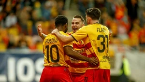 Trzech piłkarzy w żółto-czerwonych strojach drużyny świętuje zdobycie gola na murawie stadionu, obejmując się i okazując radość, w tle widać rozmyte sylwetki innych kibiców i elementy trybun.