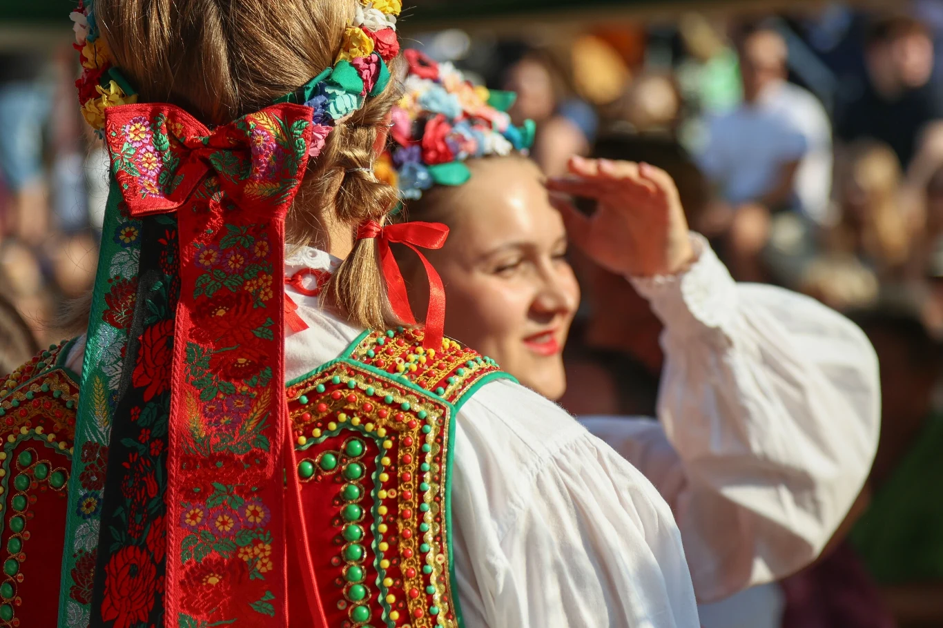 Kolorowe stroje ludowe z bogato zdobionymi elementami, jedna z kobiet zwrócona przodem zasłania oczy dłonią przed słońcem, druga widoczna z tyłu z ozdobną wstążką we włosach, w tle rozmyta grupa ludzi.