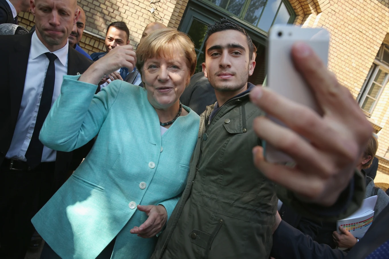 Anas Modamani i Angela Merkel w 2015 roku Kobieta ubrana w jasny żakiet i młody mężczyzna w kurtce robią sobie wspólne selfie smartfonem, wokół nich stoi kilka innych osób oraz ochroniarze, w tle widoczny budynek z żółtej cegły.