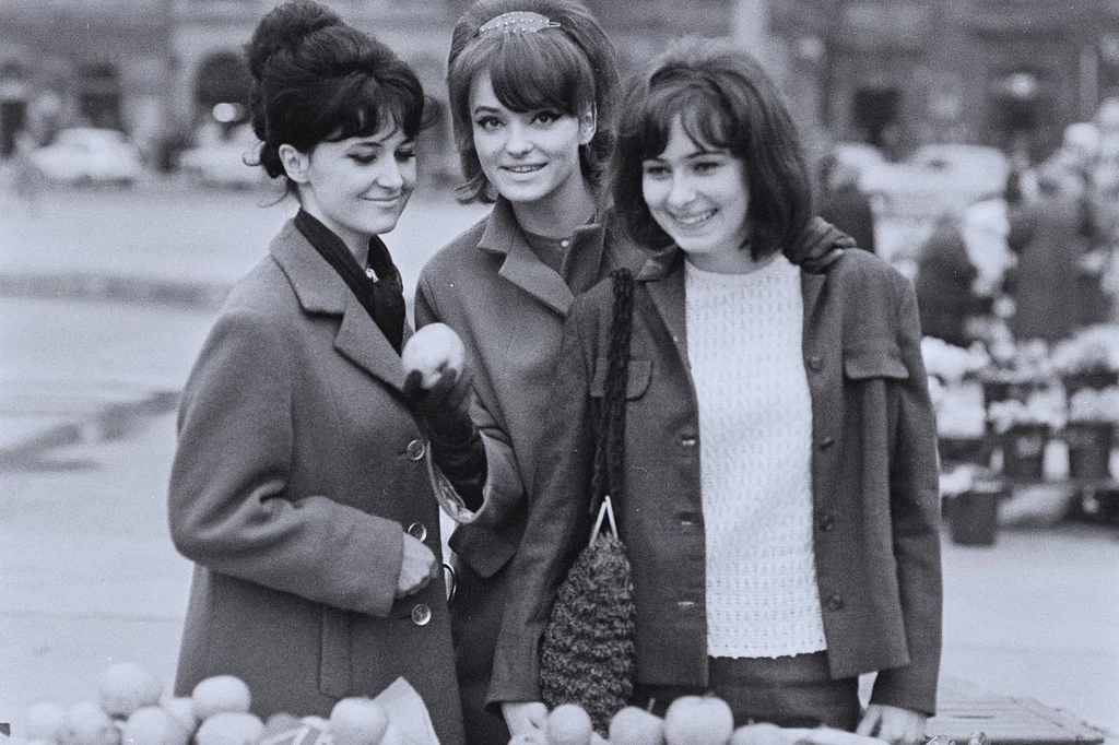 Kraków, lata 60. Trzy młode kobiety w eleganckich płaszczach stoją razem przy straganie z owocami, uśmiechnięte, w tle widoczny zarys miasta i rozmyte sylwetki samochodów.