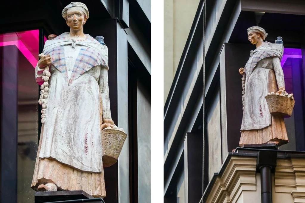 Pomnik Miedzianny, krakowskiej przekupki / Beata Zawrzel/REPORTER Rzeźba kobiety w tradycyjnym stroju z koszem, stojąca na zewnętrznej krawędzi budynku, ubrana w długą suknię z fartuszkiem i narzutką w kratę, na głowie ma czepiec, a w jednej ręce niesie wiązkę czosnku.