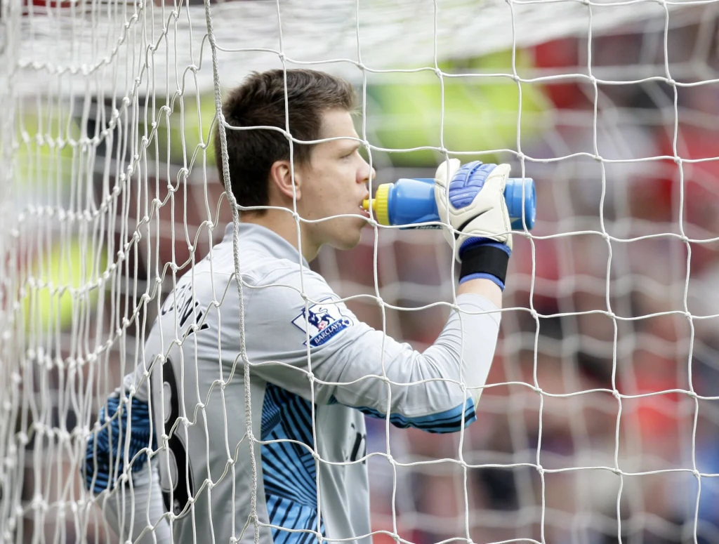 Bramkarz w jasnym stroju pije wodę z butelki, stojąc za siatką bramki na stadionie piłkarskim.