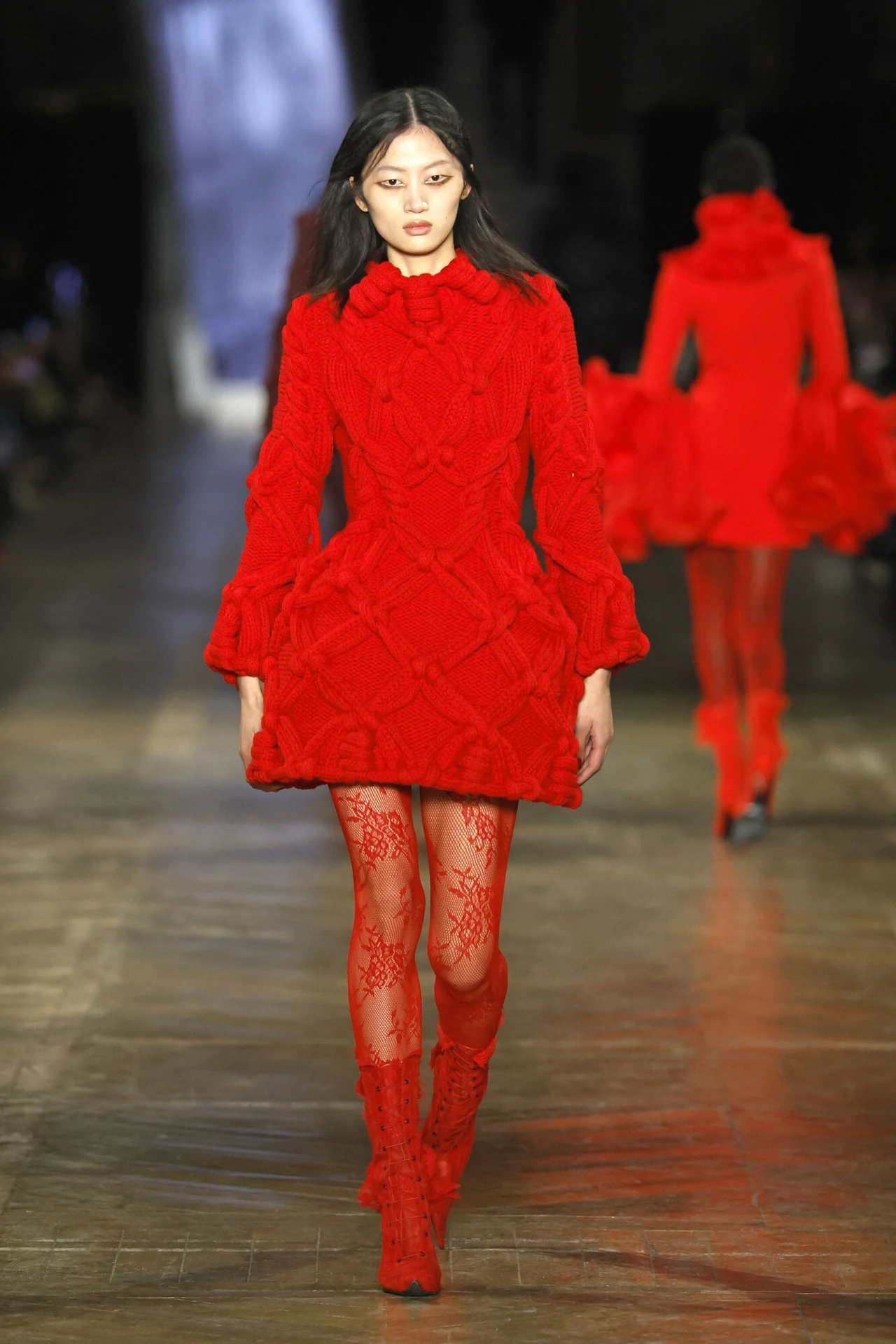 Czerwień króluje również w kolekcjach najważniejszych projektantów. Na zdj. pokaz kolekcji jesień-zima 2025/2026 Alexandra McQueena na Paris Fashion Week Modelka ubrana w efektowną, czerwoną sukienkę z bogatą fakturą oraz dopasowanymi rajstopami i butami przechodzi po wybiegu podczas pokazu mody, w tle widoczna inna osoba w podobnej stylizacji.