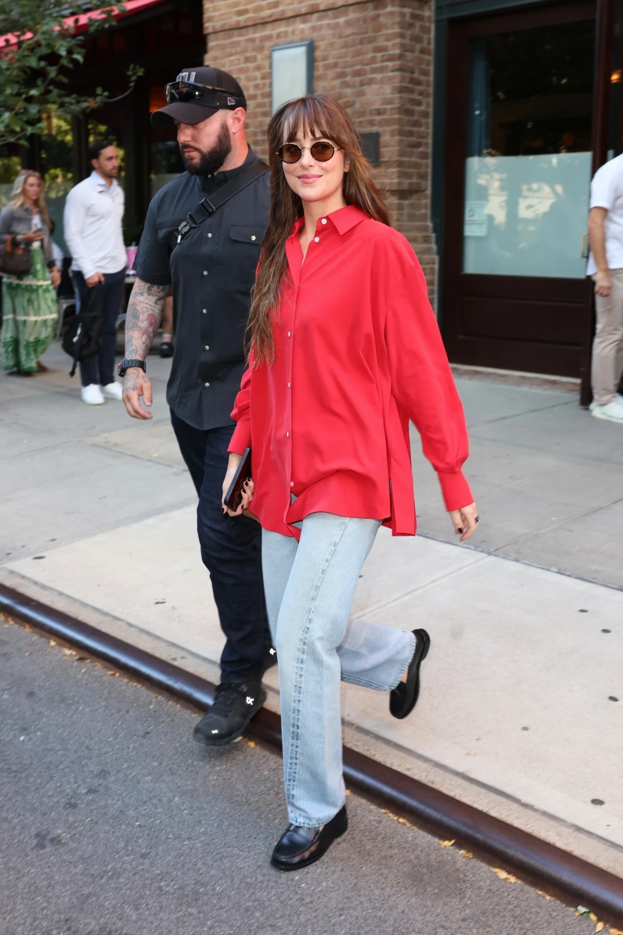 Dakota Johnson nosi od dawna. Czerwona koszula będzie hitem jesieni 2025 Kobieta w okularach przeciwsłonecznych, ubrana w czerwoną, luźną koszulę i jasnoniebieskie dżinsy spaceruje chodnikiem, obok niej idzie mężczyzna w czarnej koszuli i czapce z daszkiem; w tle widoczne osoby oraz fragmenty budynku.