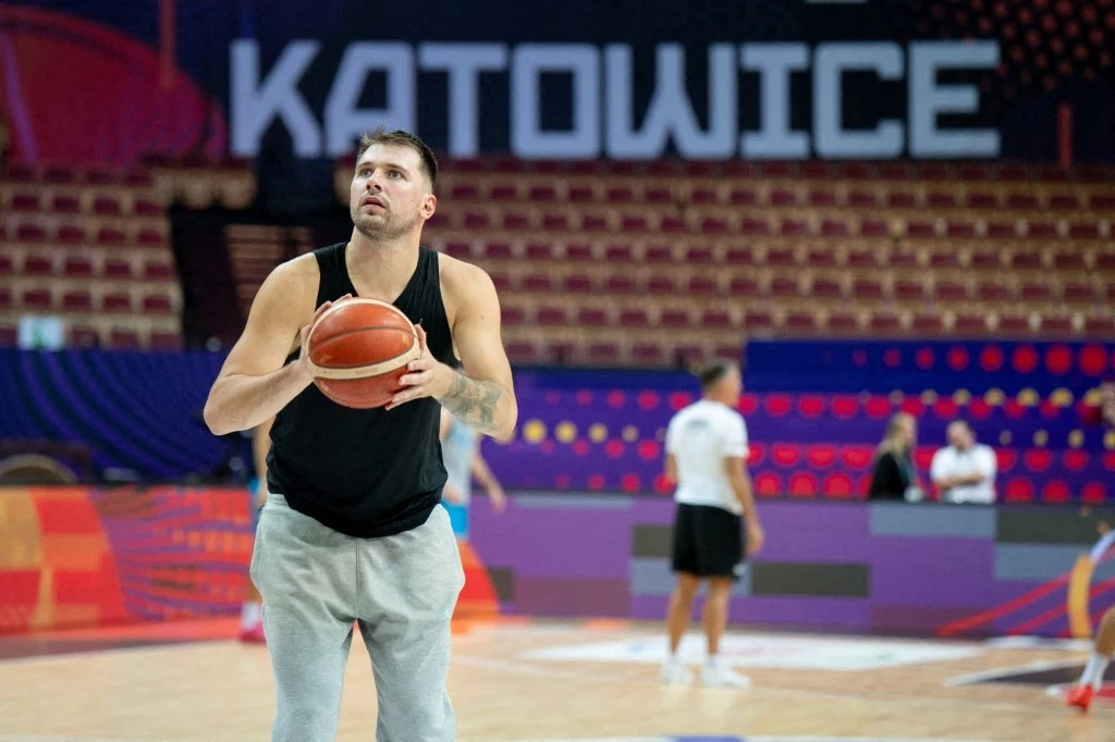 Koszykarz w czarnej koszulce i szarych spodniach szykuje się do rzutu wolnego na hali sportowej z pustymi trybunami i napisem Katowice w tle.