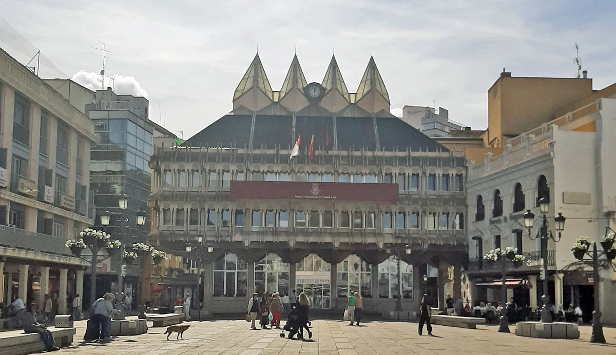 Ayuntamiento, czyli Ratusz zaprojektowany przez Fernando Higuerasa Plac otoczony budynkami z charakterystycznym, nowoczesnym gmachem w tle, na którym widoczne są flagi. Ludzie spacerują, odpoczywają i rozmawiają przy ławkach oraz stolikach ustawionych na otwartym placu.