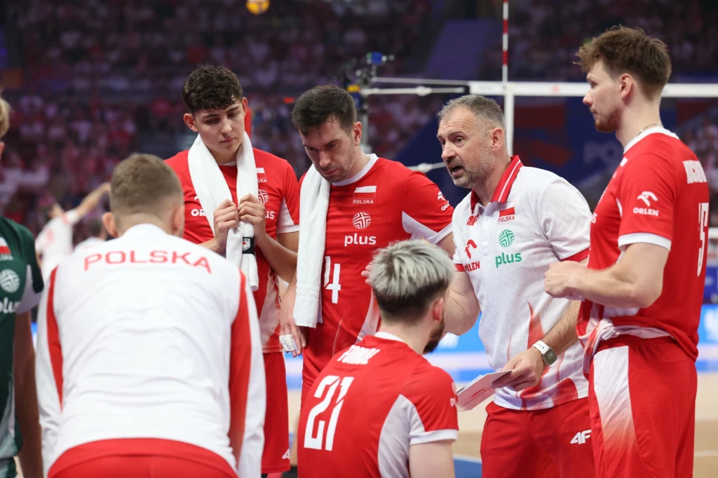 Grupa polskich siatkarzy podczas przerwy, otaczających trenera, który przekazuje wskazówki taktyczne, wszyscy w oficjalnych strojach reprezentacji, w tle rozmyte trybuny pełne kibiców.