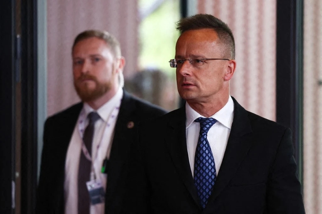 Szef węgierskiego MSZ Peter Szijjarto poinformował o zakazie wjazdu do kraju dla ukraińskiego dowódcy. Ten jest etnicznym Węgrem Dwóch mężczyzn w garniturach, jeden z nich w okularach i niebieskim krawacie, idący korytarzem, tło jest rozmyte, co skupia uwagę na osobach w pierwszym planie.