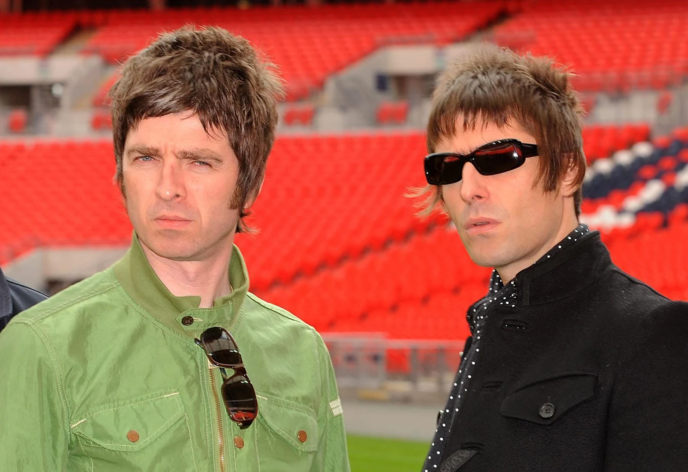 Noel i Liam Gallagher Dwóch mężczyzn stoi na tle czerwonych trybun stadionu, jeden z nich ubrany jest w zieloną kurtkę, a drugi w ciemną z polka dots, nosi ciemne okulary przeciwsłoneczne