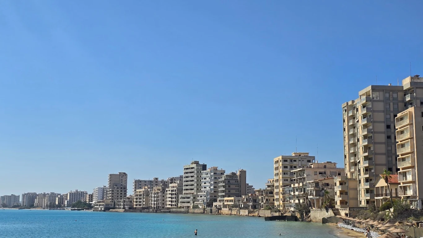 Opuszczone, wysokie budynki mieszkalne ciągnące się wzdłuż wybrzeża, pusta plaża oraz spokojne, błękitne morze pod czystym niebem