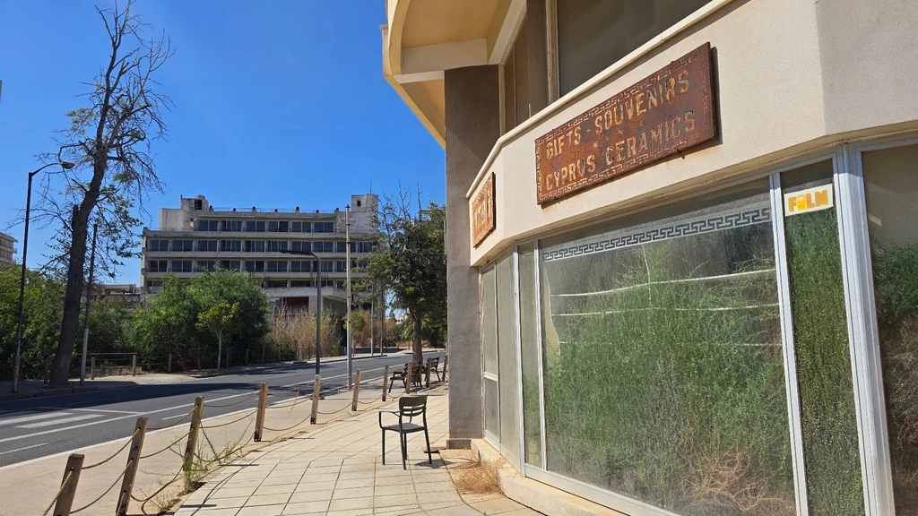 Matka natura zawładnęła przestrzenią Warosii. Opuszczony sklep z napisem „Gifts Souvenirs Cyprus Ceramics” oraz puste witryny przykryte kurzem i zarośnięte trawą, pusta ulica, po lewej stronie widoczny budynek i drzewa, atmosfera zaniedbania i opuszczenia.