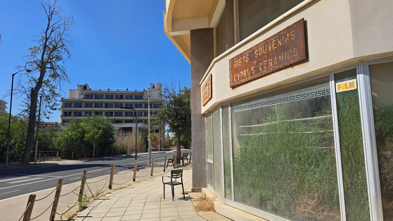 Opuszczony sklep z napisem „Gifts Souvenirs Cyprus Ceramics” oraz puste witryny przykryte kurzem i zarośnięte trawą, pusta ulica, po lewej stronie widoczny budynek i drzewa, atmosfera zaniedbania i opuszczenia.