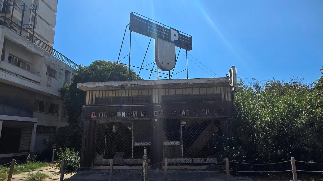 Opuszczony, zniszczony budynek z dużym, uszkodzonym szyldem parkingu na dachu, otoczony roślinnością, o fasadzie zasłoniętej kratami i wyblakłym napisem.