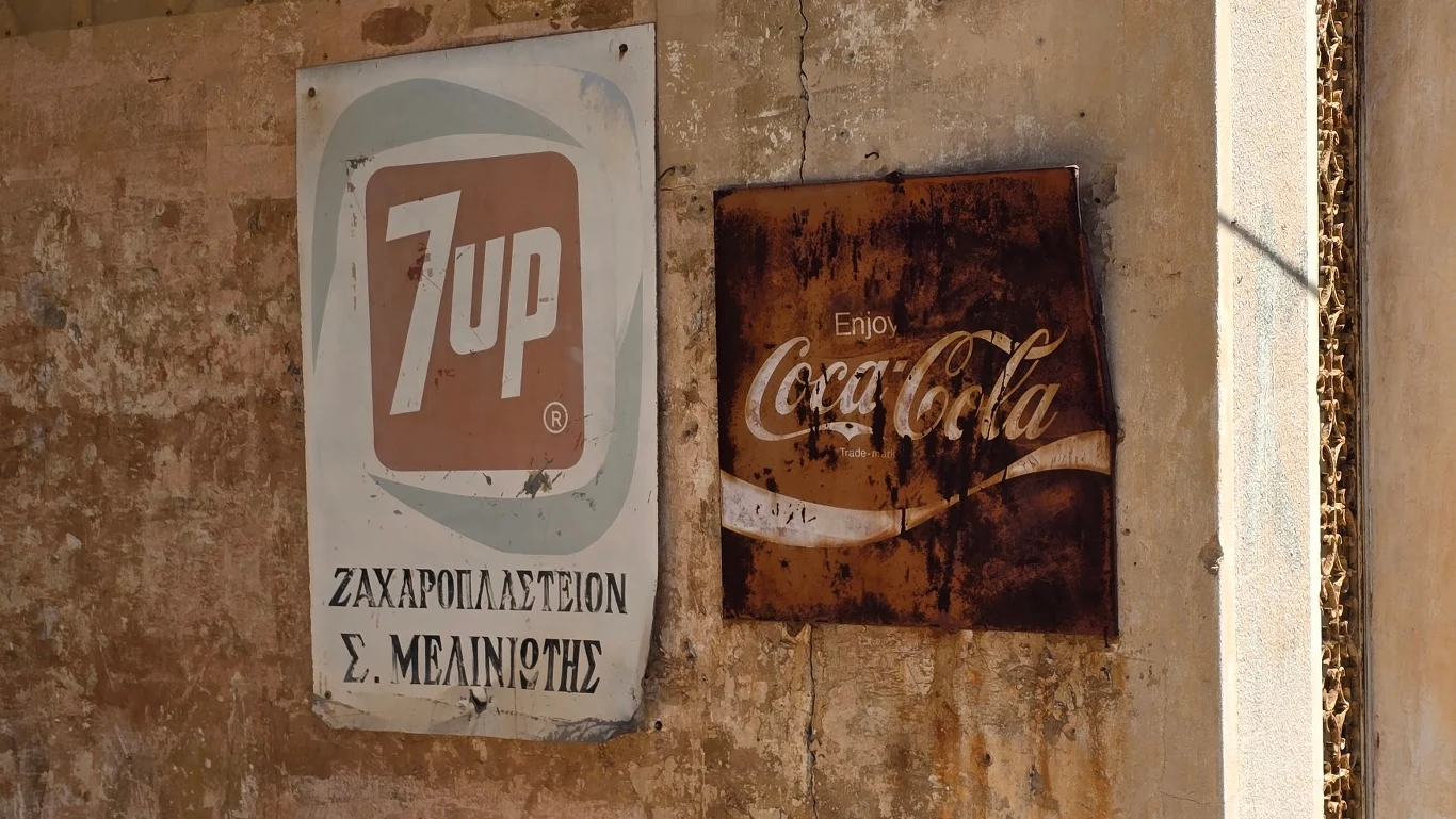 Stare, zniszczone tablice reklamowe 7up i Coca-Cola wiszą na mocno zniszczonej, brudnej ścianie budynku; tekst na tablicy 7up jest częściowo po grecku.