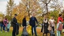 Grupa ludzi sprzątająca park w jesiennej scenerii, trzymają worki na śmieci, w tle widoczne kolorowe liście na drzewach.