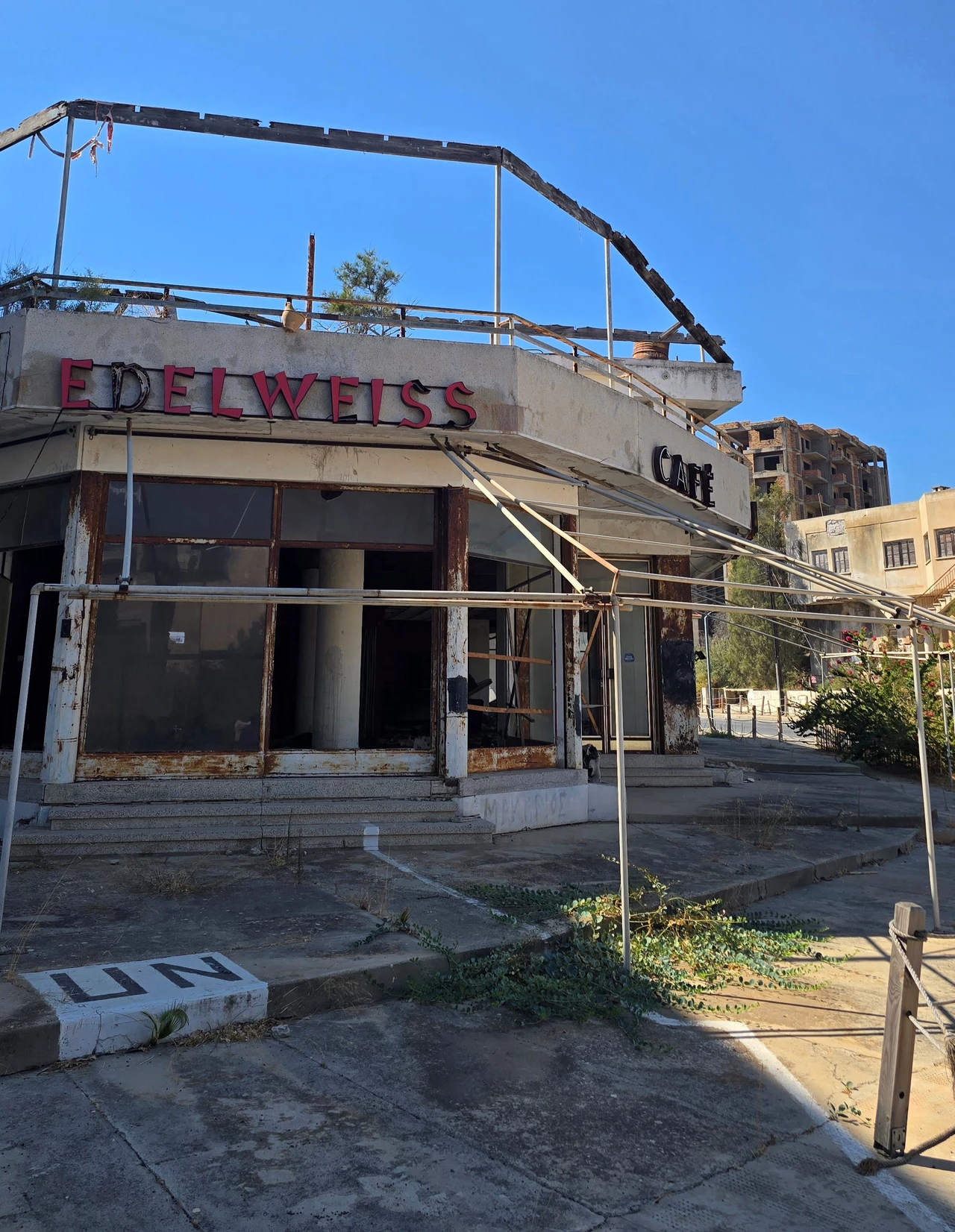 Edelweiss Cafe w centrum Warosii. Tuż obok znajduje się punkt Organizacji Narodów Zjednoczonych (UN). Zniszczony, opuszczony budynek kawiarni z widocznym napisem 'EDELWEISS CAFE', skażone szybami i zardzewiałą konstrukcją w tle, wokół obiektu zaniedbana roślinność i betonowy chodnik, w tle inne stare budynki pod niebieskim niebem.
