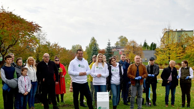 Grupa ludzi różnych wiekiem i płci, stojących na zielonej trawie otoczonej jesiennymi drzewami podczas wspólnej akcji sadzenia drzew, z tabliczką kampanii ekologicznej na pierwszym planie.