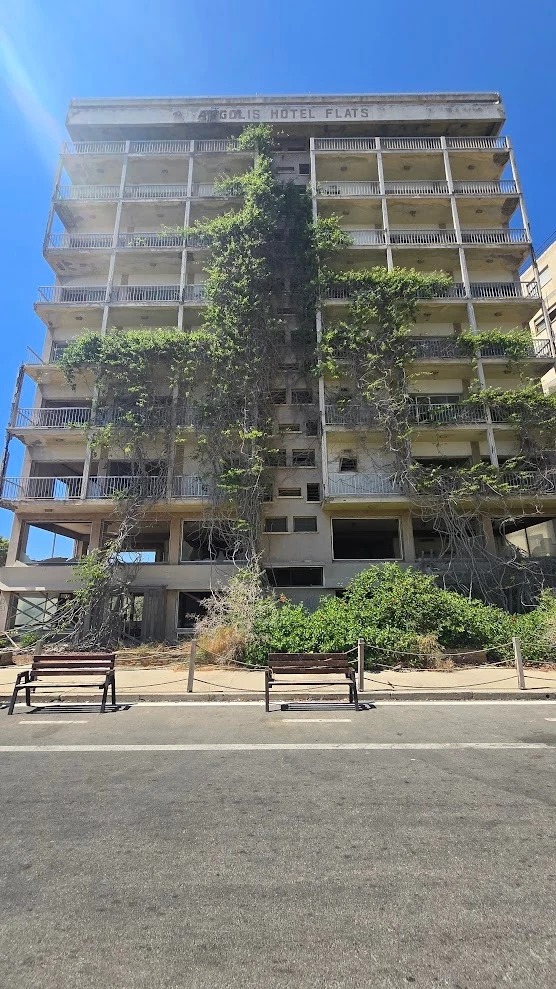 Wielokondygnacyjny opuszczony budynek hotelowy porośnięty bujną zielenią oraz winoroślami wspinającymi się po jego fasadzie. Na pierwszym planie dwa puste drewniane ławki oraz ogrodzenie oddzielające chodnik od wejścia do hotelu. Jasne niebo bez chmur ...