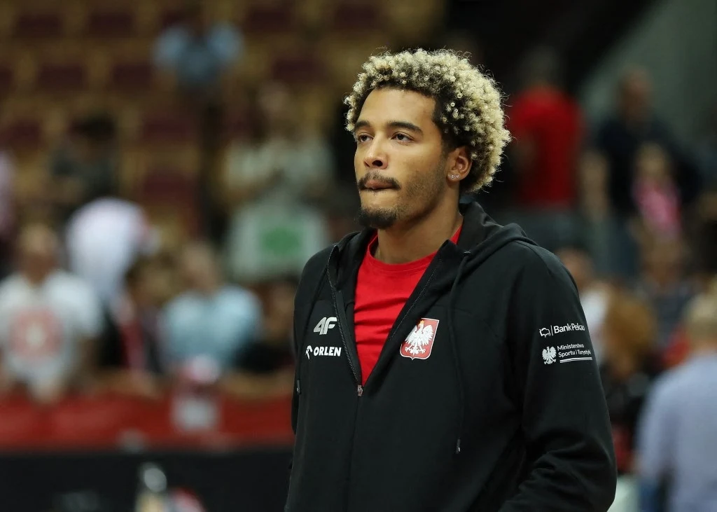 Młody mężczyzna w sportowej bluzie z orłem na piersi stoi na tle zamazanej widowni, skupiony i poważny, ubrany w czarno-czerwony strój reprezentacyjny.