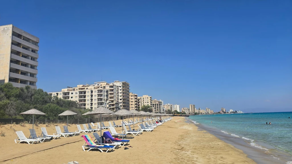 Plaża w mieście duchów na Cyprze. Współczesne leżaki i ruiny budynków, w których hula wiatr. Pusta, piaszczysta plaża z rzędami leżaków i parasoli, w tle opuszczone, wysokie budynki przy linii brzegu oraz lazurowe morze pod bezchmurnym niebem.