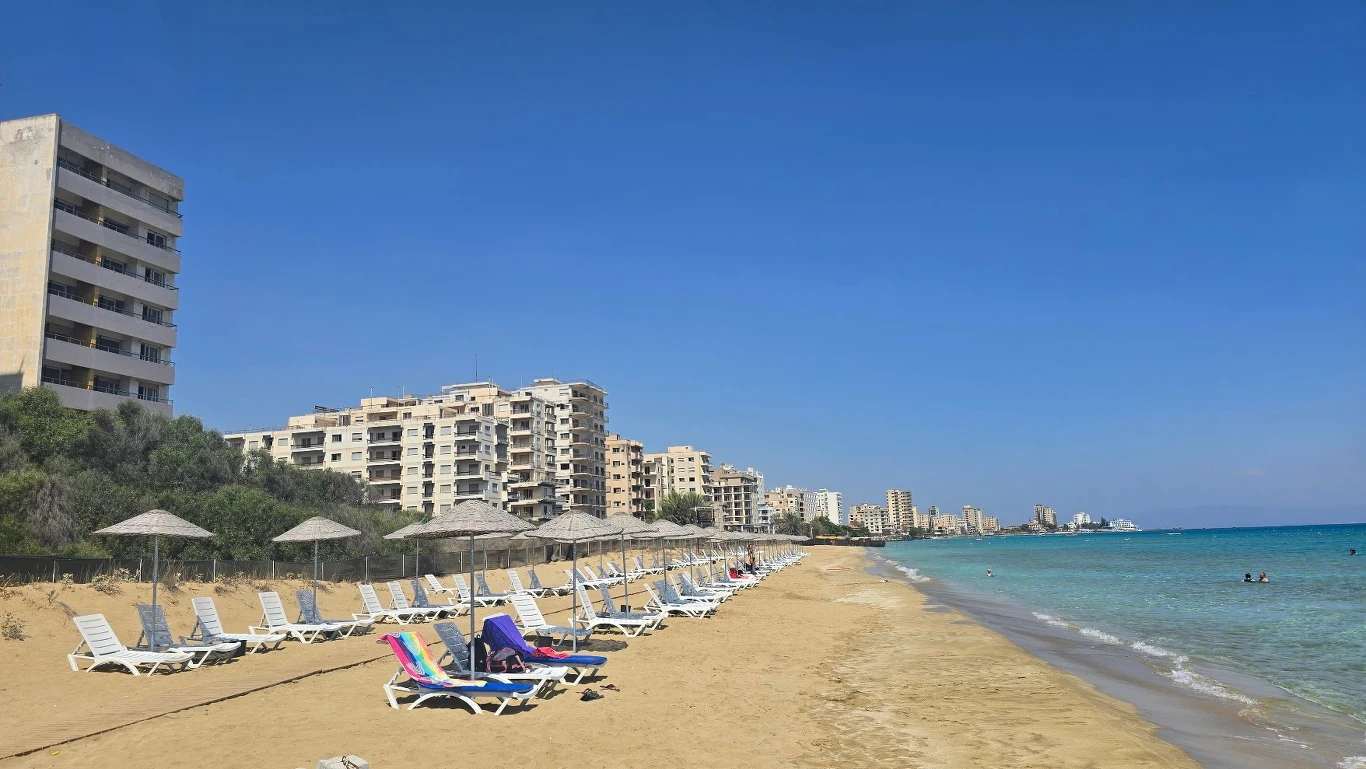 Pusta, piaszczysta plaża z rzędami leżaków i parasoli, w tle opuszczone, wysokie budynki przy linii brzegu oraz lazurowe morze pod bezchmurnym niebem.