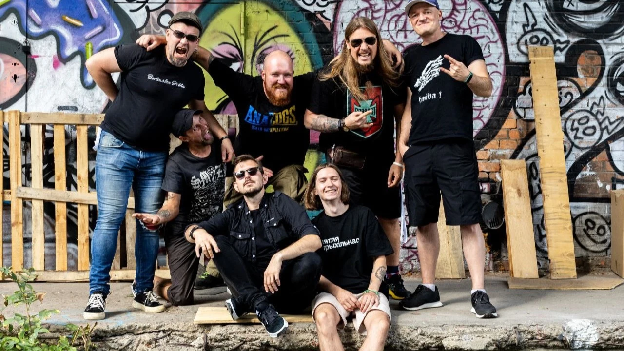 Zespół Dzieciuki Grupa siedmiu uśmiechniętych mężczyzn pozuje razem przy ścianie pokrytej kolorowymi graffiti, część z nich stoi, a część siedzi na krawędzi betonowej powierzchni, w tle znajdują się palety i drewniane elementy.