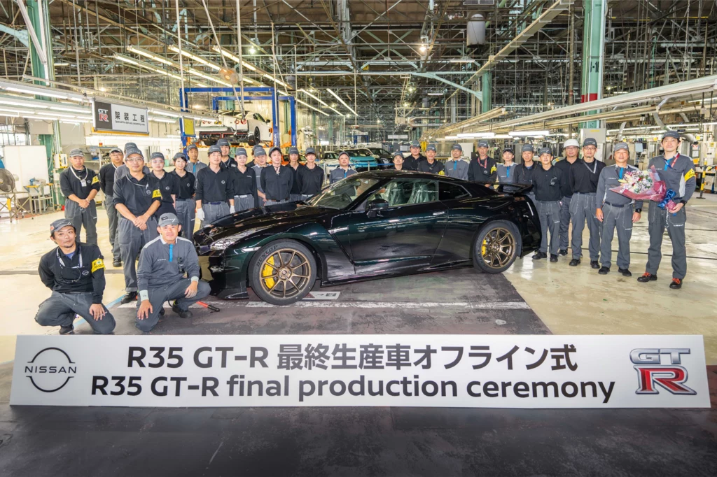 Czarny samochód sportowy Nissan GT-R R35 otoczony przez zespół pracowników fabryki w hali produkcyjnej, na pierwszym planie widoczny baner informujący o ceremonii zakończenia produkcji tego modelu.