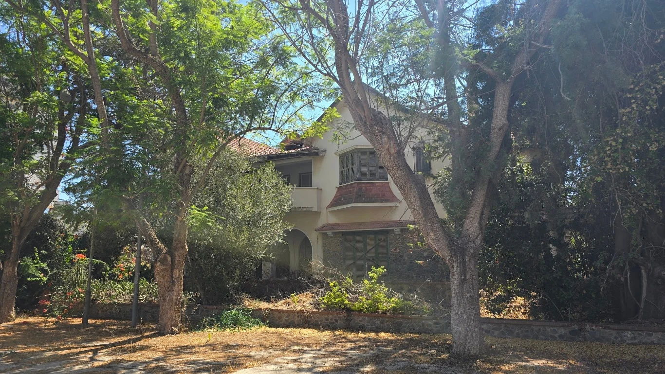 Dwupiętrowy dom o jasnych ścianach skryty wśród wysokich, liściastych drzew, z czerwonym dachem i widocznymi łukami przy wejściu, otoczony gęstą roślinnością i suchymi gałęziami.