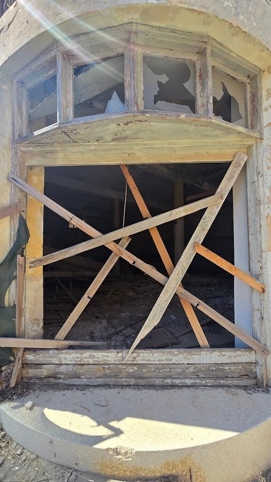 Zniszczona, zabita deskami okienna wnęka w opuszczonym budynku, ze stłuczonymi szybami i widocznymi śladami dewastacji oraz kurzem i gruzem wokół otworu.