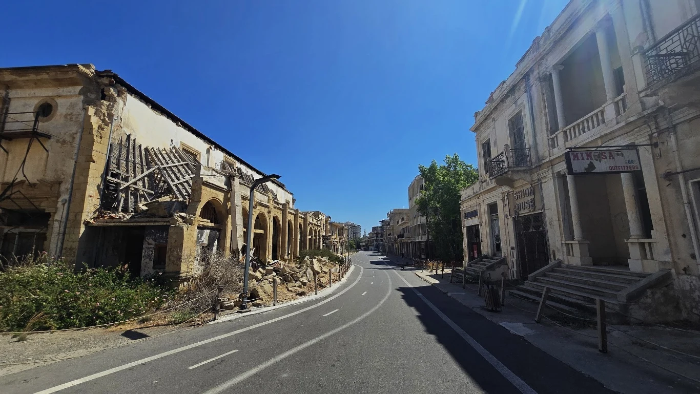 Opuszczona ulica otoczona zrujnowanymi budynkami, z których niektóre mają uszkodzone fasady i zawalone dachy, cały teren wydaje się opustoszały i zaniedbany, pośród upalnej pogody i bez obecności ludzi.