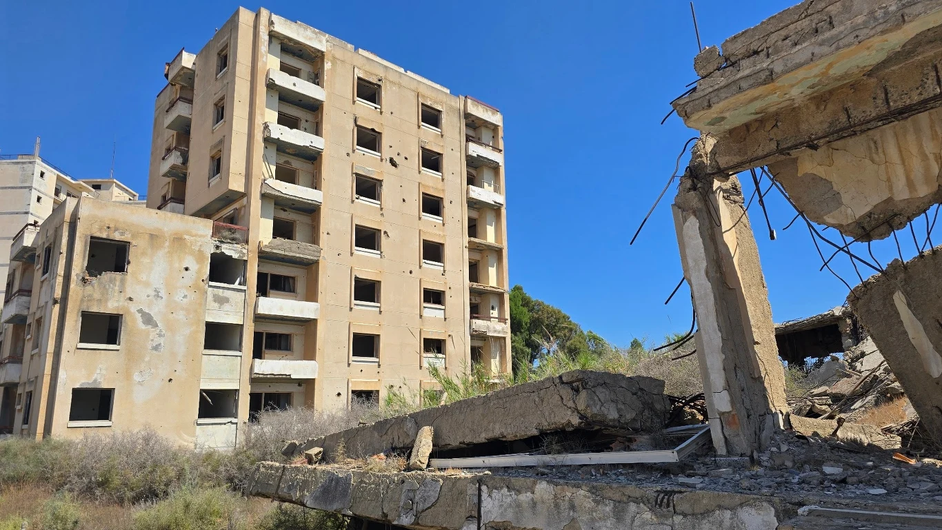 Zniszczony wielopiętrowy budynek mieszkalny z licznymi śladami uszkodzeń, fragmentem zawalonej konstrukcji na pierwszym planie, otoczony suchą roślinnością pod błękitnym niebem.