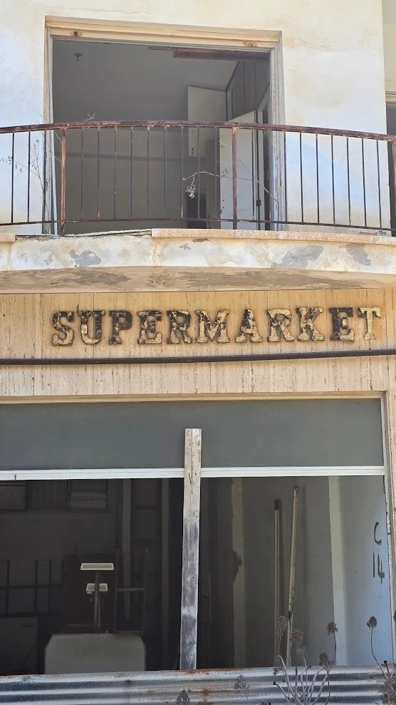 Opuszczony budynek supermarketu z widocznym zniszczonym szyldem, zaniedbanymi oknami oraz pozostałościami wyposażenia w środku.