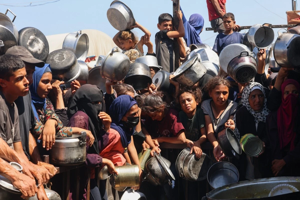 Dramatyczna sytuacja humanitarna w Strefie Gazy Tłum ludzi, głównie kobiet i dzieci, trzymających metalowe garnki i naczynia, stojący w kolejce w oczekiwaniu na jedzenie, wyrażający zmęczenie i niepokój.