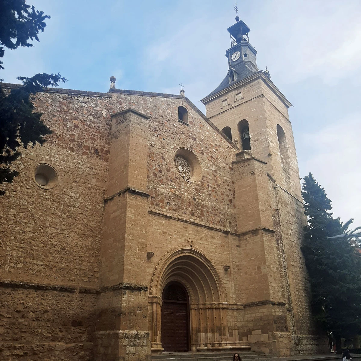 Kościół Sw Piotra w Ciudad Real kamienny kościół z wysoką wieżą zegarową, masywnymi filarami i ozdobionym wejściem z łukiem, wokół kościoła rosną drzewa, a cała budowla dominuje nad otoczeniem