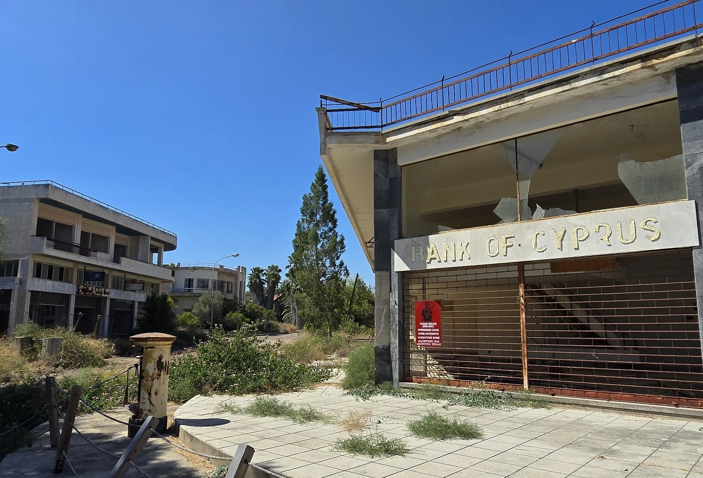 Opuszczony budynek banku z zamkniętą kratą oraz sąsiednie zaniedbane zabudowania otoczone przez roślinność, scena pustki i zaniedbania w mieście.