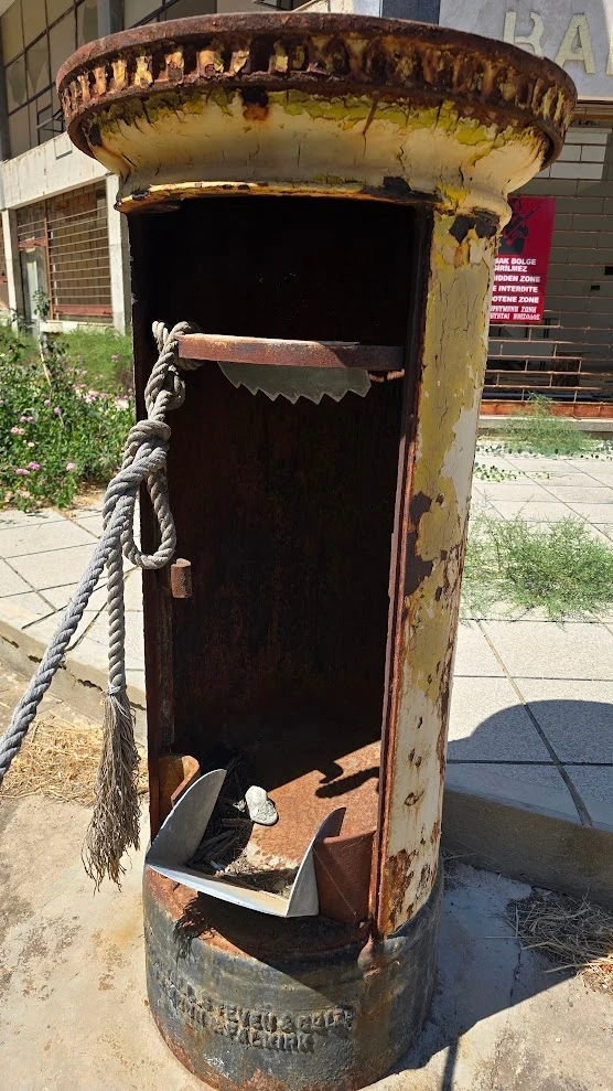 Zardzewiała, opuszczona skrzynka pocztowa o żółtej farbie złuszczonej z powierzchni, z wnętrzem wyeksponowanym po usunięciu drzwiczek, przewiązana grubym sznurem; w środku widoczne metalowe elementy oraz odpadki, otoczenie z betonowymi płytami i chwast...