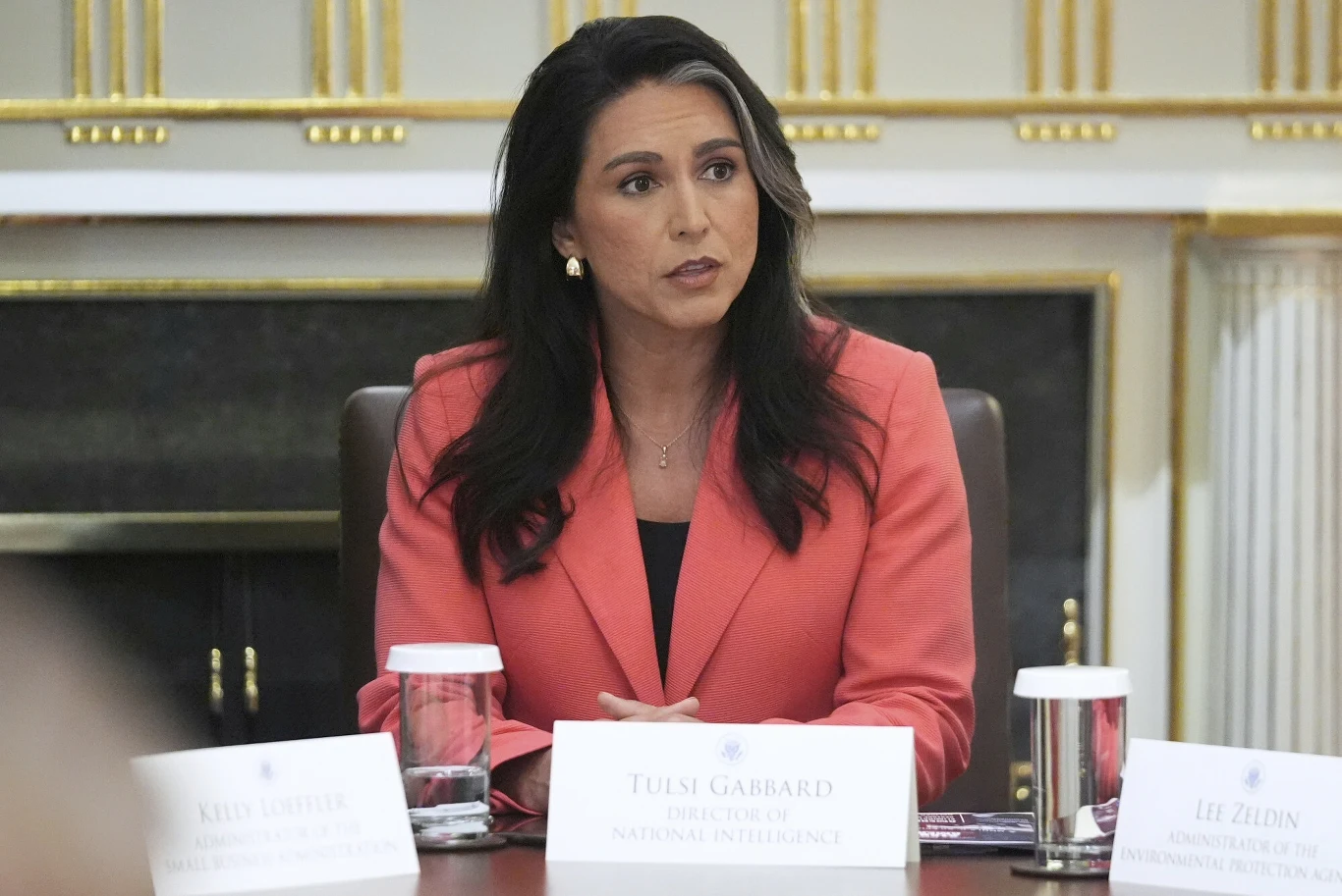 Szefowa Wywiadu Narodowego USA Tulsi Gabbard Kobieta w czerwonej marynarce siedzi przy stole konferencyjnym, przed nią leży plakietka z nazwiskiem, w tle eleganckie wnętrze z pozłacanymi detalami, po bokach widoczne są papierowe kubki i nazwiska innych uczestników spotkania.