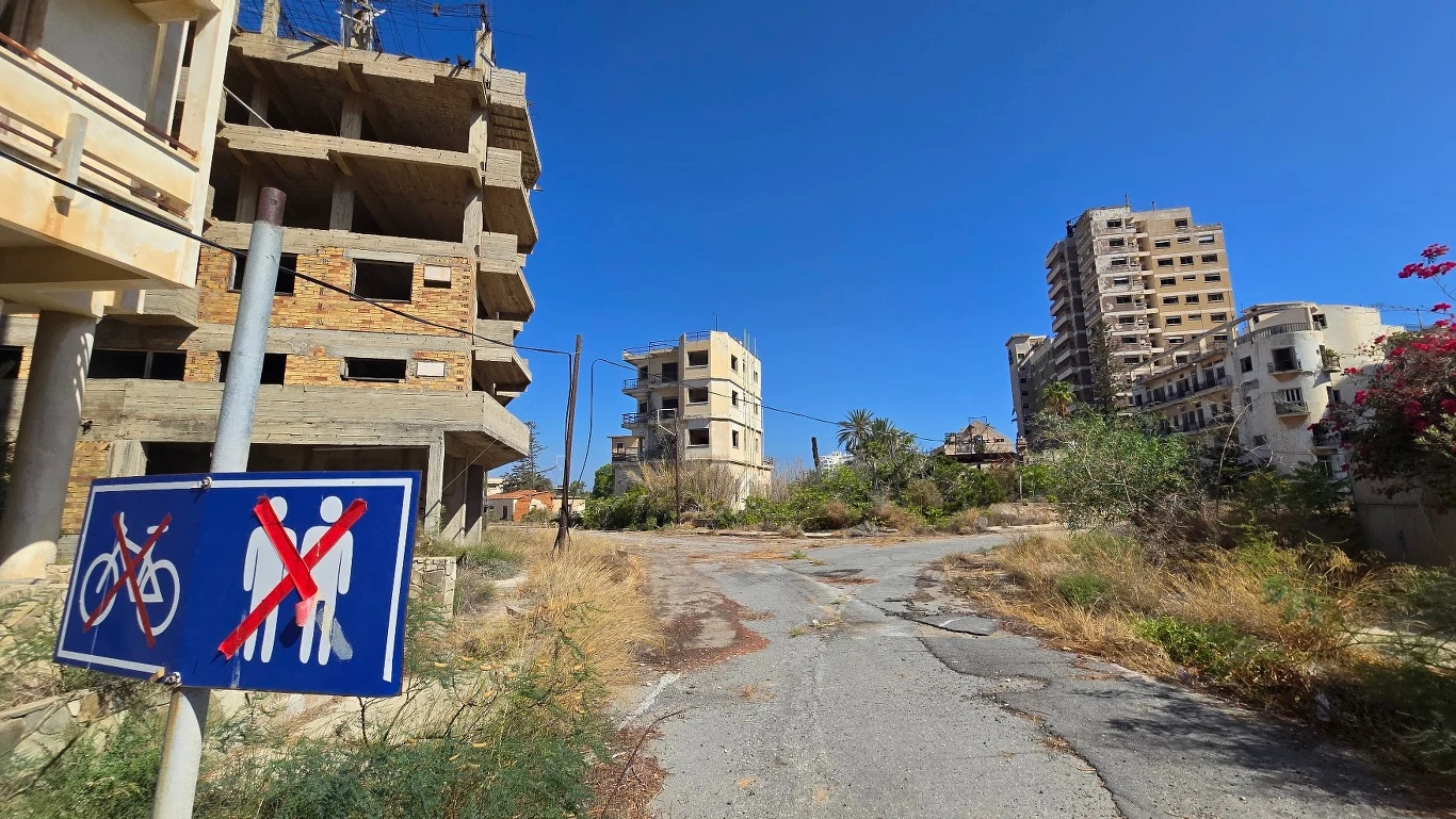 Opuszczone, częściowo zniszczone budynki przy zaniedbanej ulicy, znak z zakazem wstępu dla pieszych i rowerzystów, wokół porośnięta roślinność i brak ludzi.