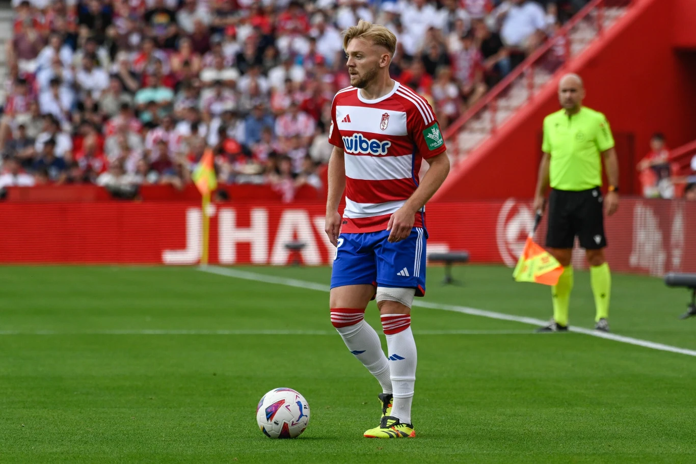 Kamil Jóźwiak Piłkarz w stroju drużyny Granada CF stoi na murawie stadionu z piłką przy nodze, w tle widać sędziego oraz trybuny wypełnione kibicami.