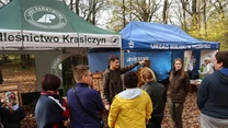 Grupa ludzi zgromadzona pod namiotami Lasów Państwowych i Urzędu Miejskiego, otoczona jesiennymi liśćmi w parku, uczestniczy w rozmowie z osobami ubranymi w leśnicze mundury.