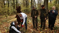 Grupa osób sadzi drzewka w lesie podczas jesieni, kilku mężczyzn i kobieta uczestniczy w akcji, obok znajduje się osoba przebrana za misia oraz mężczyzna robiący zdjęcie telefonem.