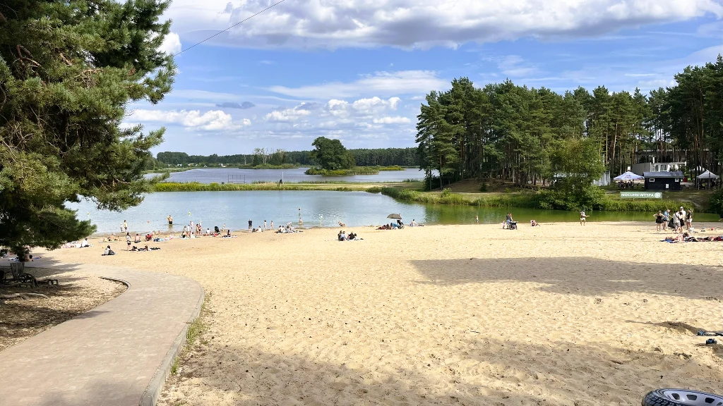 Plaża z żółtym piaskiem otoczona drzewami, kąpielisko z osobami wypoczywającymi nad wodą, na drugim planie widoczne jezioro oraz budynki wśród lasu, krajobraz letni z lekkim zachmurzeniem na niebie.