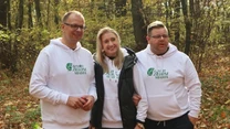 Trójka dorosłych w białych bluzach z napisem „Czyste Zielone Miasta” stoi wspólnie w jesiennym lesie, otoczeni opadłymi liśćmi i drzewami.