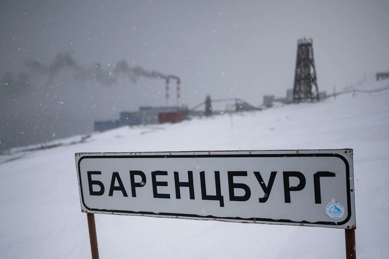 Witamy w Barentsburgu Tablica z napisem 'Баренцбург' ustawiona na tle ośnieżonego, industrialnego pejzażu z widocznymi kominami oraz budynkami przemysłowymi, w zimowej scenerii z padającym śniegiem.