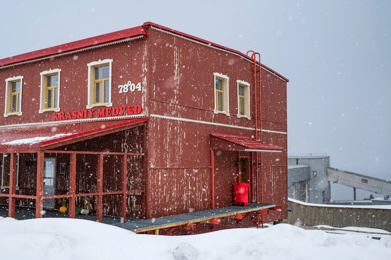 Jeden z budynków w Barentsburgu Czerwony, drewniany budynek z napisem Krasniy Medved, o numerze 78041, stojący w śnieżnej scenerii z padającym śniegiem, z wyraźnie widocznym balkonem i fragmentem innego budynku w tle.