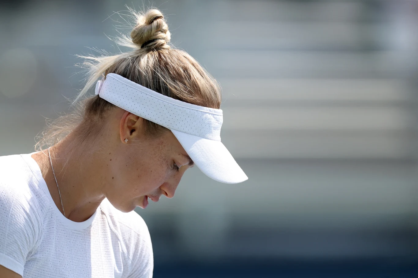 Sportowa zawodniczka w białej koszulce i białej opasce na głowie pochylona w zamyśleniu, skupiona na tle rozmytego stadionu.
