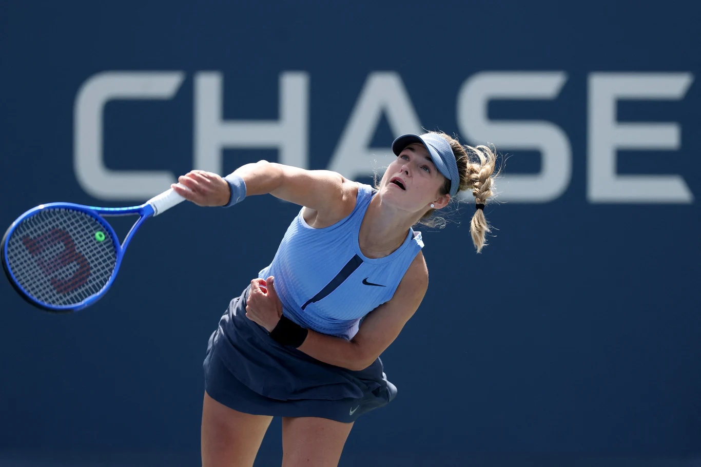 Tenisistka w niebieskim stroju tuż po wykonaniu serwisu podczas meczu na tle niebieskiego kortu i dużego białego napisu na ścianie.