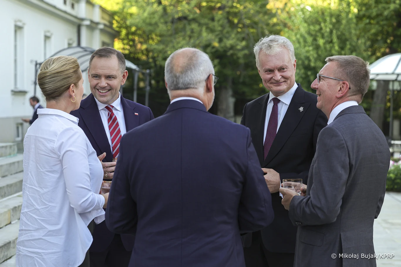 Liderzy państw, którzy zjechali do Warszawy na zaproszenie Karola Nawrockiego Grupa pięciu elegancko ubranych osób w garniturach i marynarce rozmawia i uśmiecha się, stojąc na zewnątrz przy jasnym budynku w otoczeniu zieleni, trzymając w dłoniach szklanki z napojami.