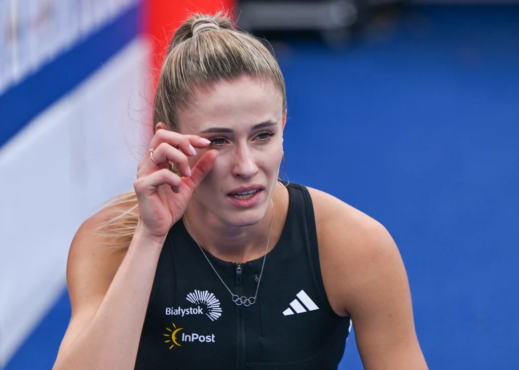 kobieta w sportowym stroju z widocznymi logotypami sponsorów trzyma rękę blisko oka, jej wyraz twarzy wskazuje na zmęczenie lub silne emocje po intensywnym wysiłku fizycznym, w tle rozmyte elementy toru sportowego