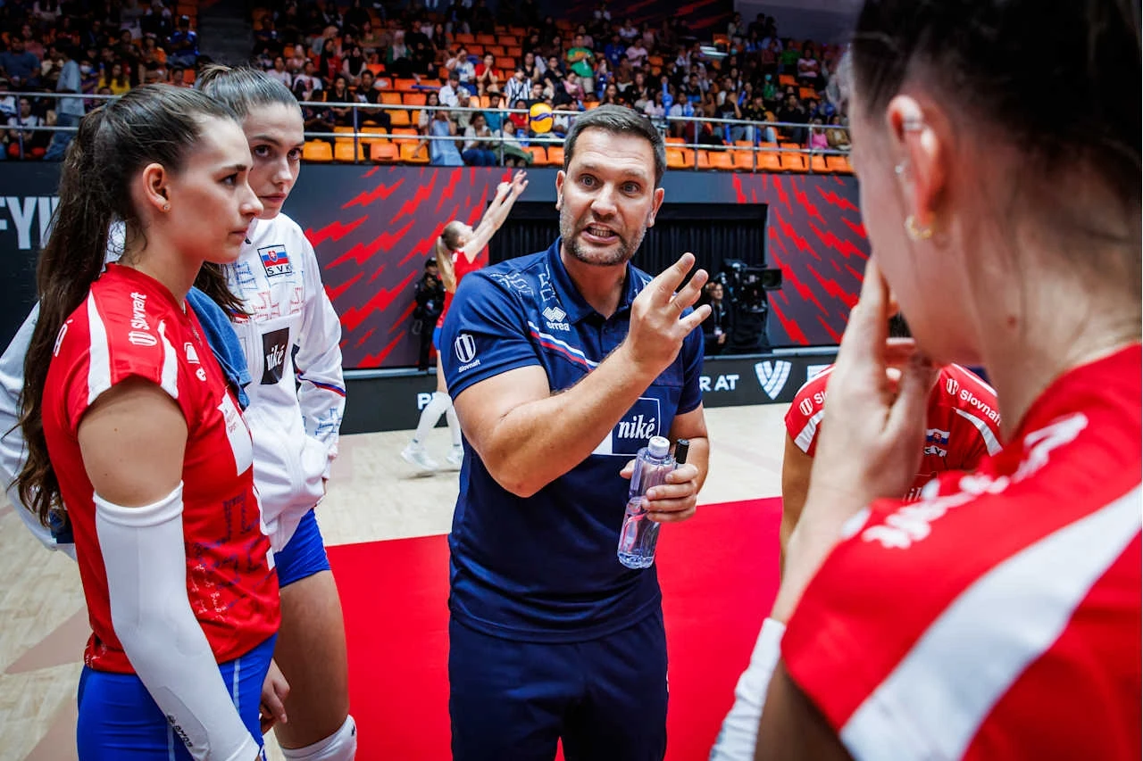 Trener siatkarski udziela wskazówek zawodniczkom swojej drużyny podczas przerwy w meczu, na pierwszym planie widoczne skupienie i zaangażowanie uczestników, w tle trybuny z kibicami.